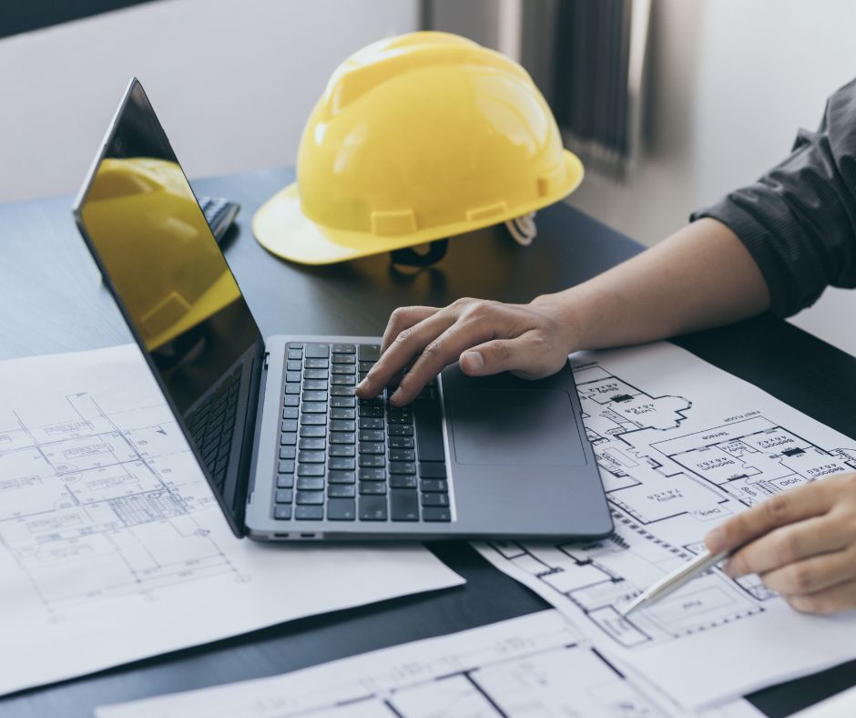 Μηχανικός δουλεύει σε laptop πάνω σε αρχιτεκτονικά σχέδια, δίπλα σε κίτρινο κράνος